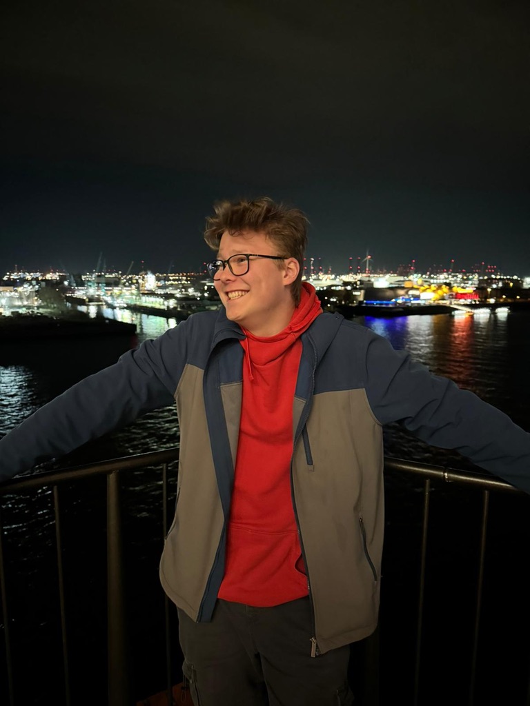 Max lacht nachts an einem Geländer, im Hintergrund ein beleuchteter Hafen